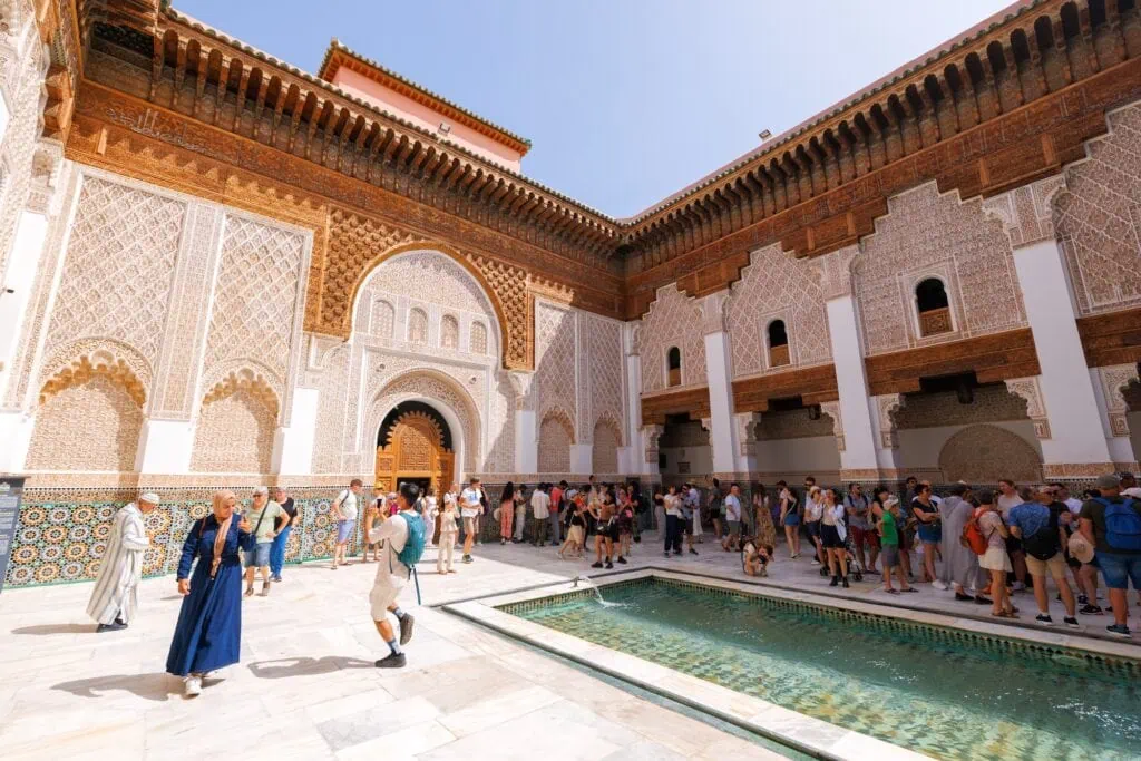 Madrasa Ben Youssef courtyard with carved stucco walls, geometric tilework, and visitors walking through the historic complex Marrakech