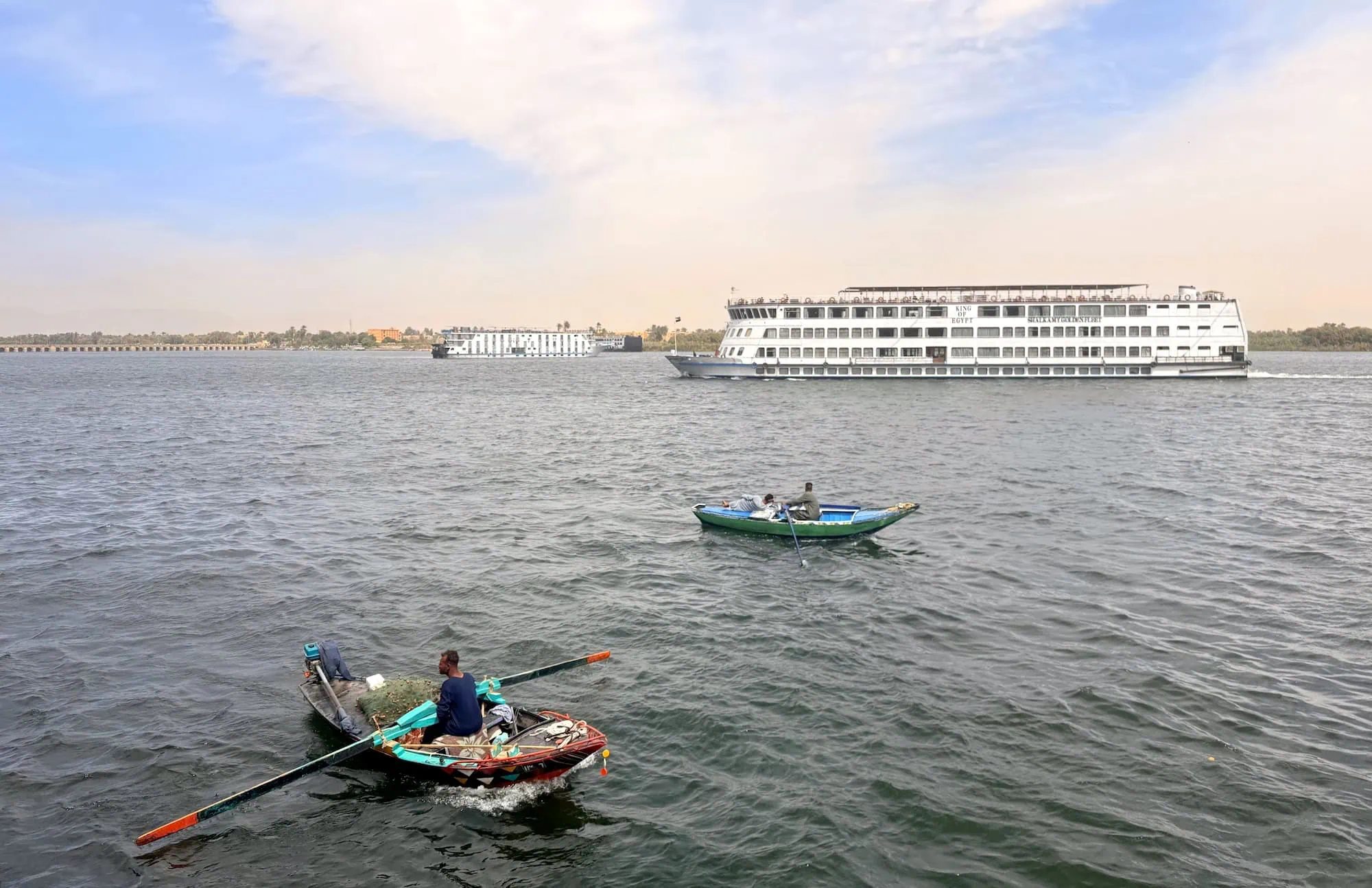 A large white multi-deck Luxury Nile Cruise vessel sailing on the Nile between Luxor and Aswan, with small traditional wooden fishing boats in the foreground and palm-lined banks behind.