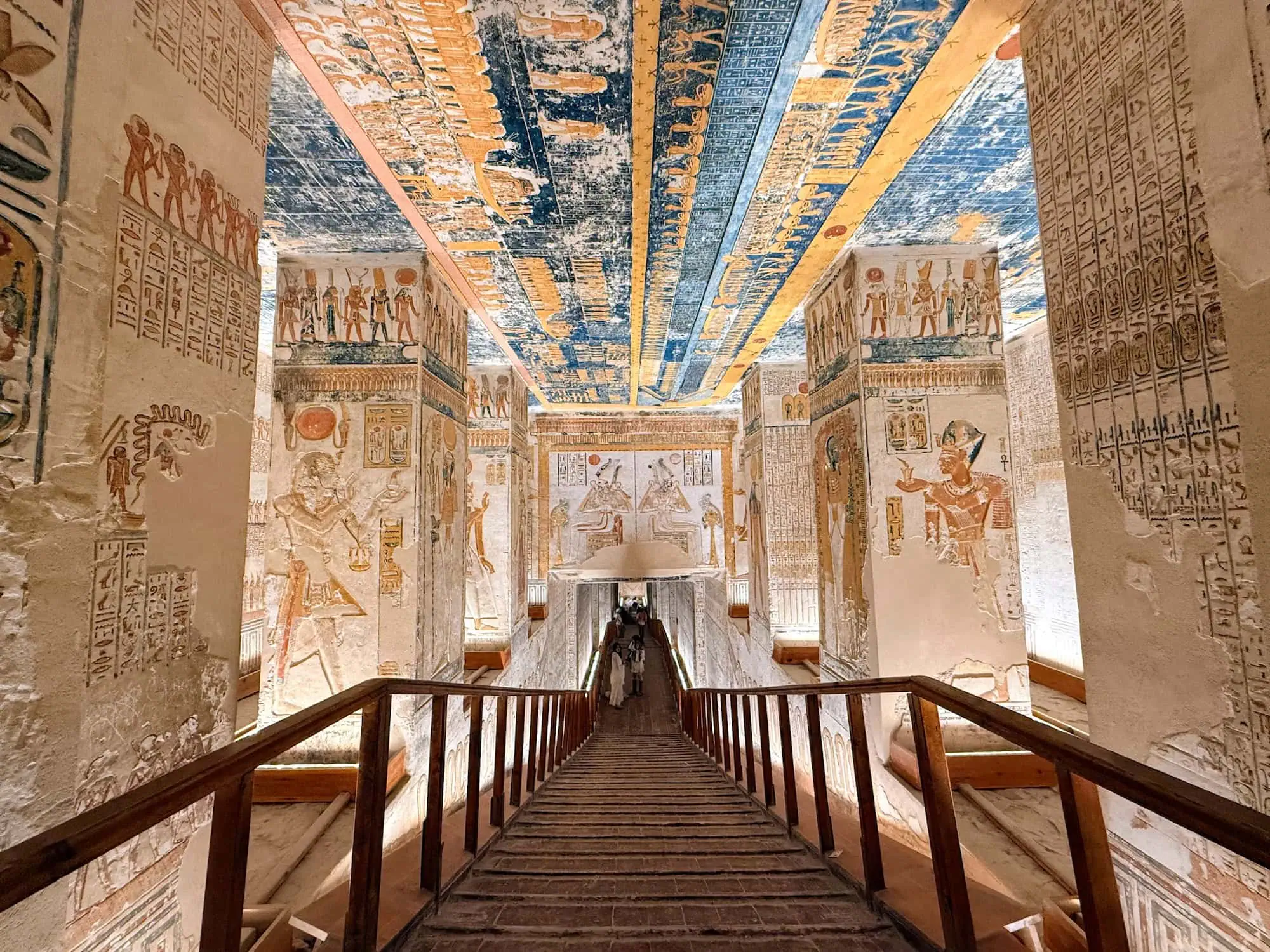 Wooden walkway descending into a pillared royal tomb in the Valley of the Kings near Luxor, with a blue-and-gold astronomical ceiling depicting the sky goddess Nut and painted reliefs of the pharaoh before the gods, visited on a guided Egypt all-inclusive Nile cruise.